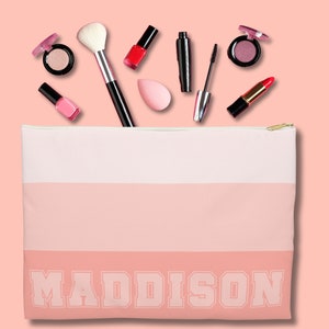 May include: A blush-toned makeup bag with the name "MADDISON" in bold, white letters. The bag is filled with makeup products, including eyeshadow, lipstick, mascara, and a makeup brush. The background is a soft pink.