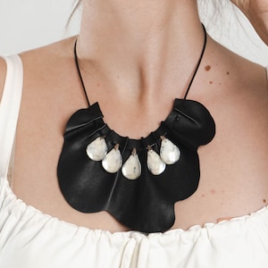 May include: A black leather necklace with a unique, ruffled design. The necklace features six teardrop-shaped, white and gray pendants suspended from the leather. The necklace is worn against a white top.