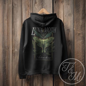 Op de afbeelding: Zwarte hoodie met een groen Luna mot ontwerp, varen accenten en de tekst "LUNA MOTH". De tekst "Actias luna" en "Ephemeral nocturne of the forest" staan eronder. De hoodie hangt aan een houten hanger.