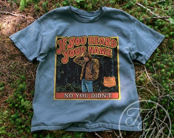 May include: A slate blue short-sleeve t-shirt featuring a graphic print of a person in a forest with a backpack. The text reads IF YOU HEARD YOUR NAME NO YOU DIDN'T, with a small badge labeled PARK SAFETY GUIDE in the corner of the square design.