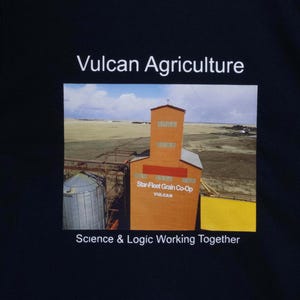 May include: Black t-shirt featuring the text "Vulcan Agriculture" above a colorful image of an orange grain elevator and the text "Science & Logic Working Together". The shirt is hanging on a black hanger.