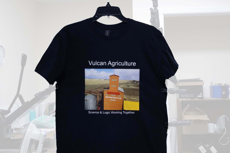 May include: Black t-shirt featuring the text "Vulcan Agriculture" above a colorful image of an orange grain elevator and the text "Science & Logic Working Together". The shirt is hanging on a black hanger.