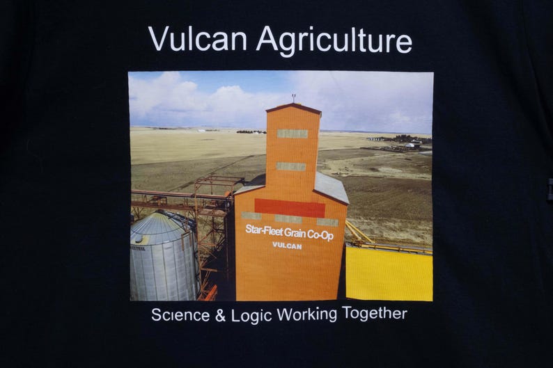 May include: Black t-shirt featuring a landscape image of an orange grain elevator with the text "Vulcan Agriculture" and "Star-Fleet Grain Co-Op". The phrase "Science & Logic Working Together" is also displayed.