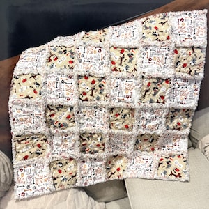 May include: A patchwork dog-themed blanket with a cream-colored background and squares featuring dog illustrations, bones, and the words "woof" and "paws." The blanket has a fringed edge and is likely made of soft fabric.
