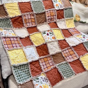 May include: A rag quilt featuring a patchwork of square fabric blocks in shades of orange, yellow, brown, and green. The quilt includes solid colors, plaid patterns, and floral prints with exposed frayed seams between each square block.