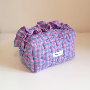 May include: A pink and blue gingham patterned quilted cosmetic bag. The bag features a frilled top and a zip closure. A small white label with the text "bykatemerris" is attached to the side.