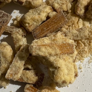 May include: A close-up of a pile of crispy, sugar-coated fried snacks. The snacks are light brown in color and have a grainy texture.