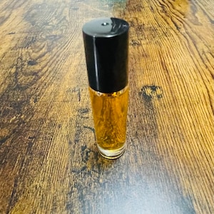 May include: A small, clear glass bottle with a black cap filled with a golden liquid. The bottle is sitting on a wooden surface.