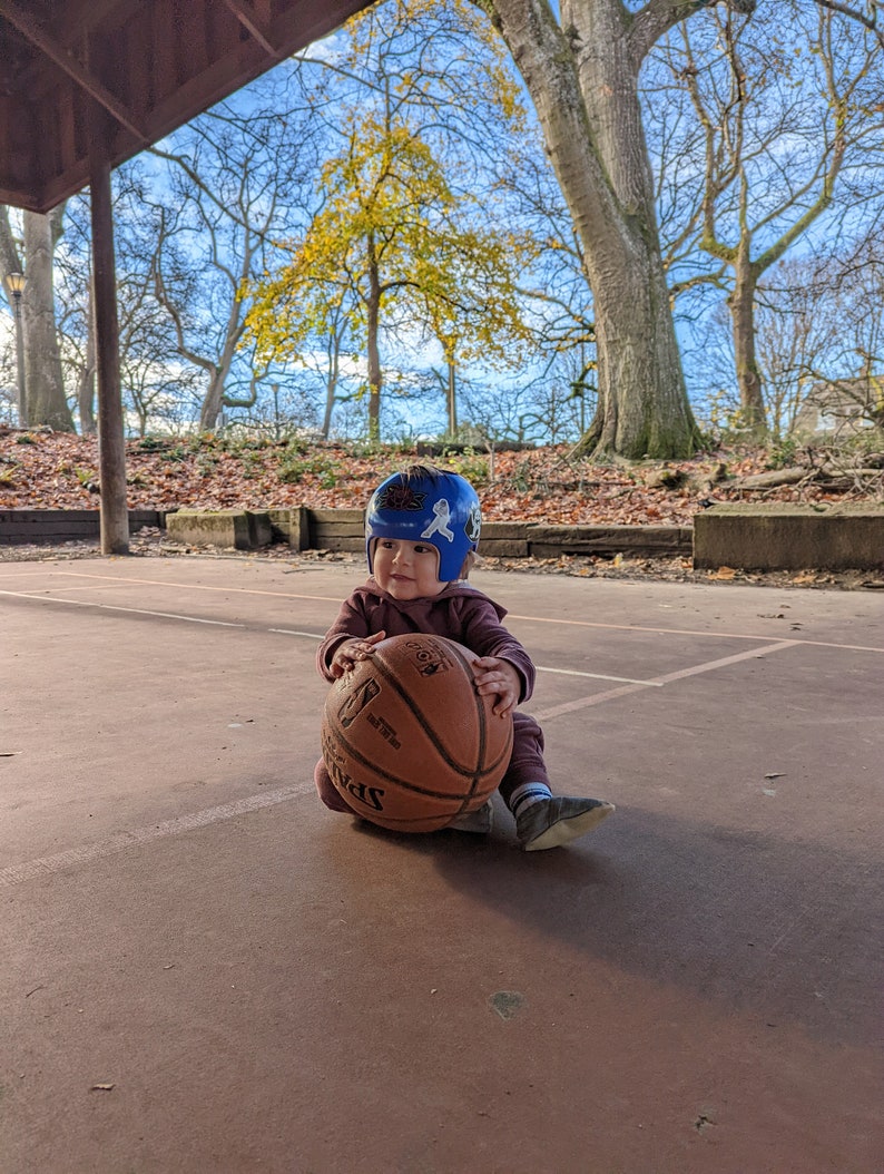 May include: A toddler wearing a blue helmet with stickers sits on a basketball court holding a brown basketball. The basketball has white markings and the word " Spalding" printed on it.