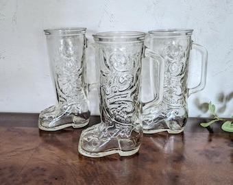 May include: Three clear glass boot-shaped mugs with handles. The mugs are on a wooden surface.