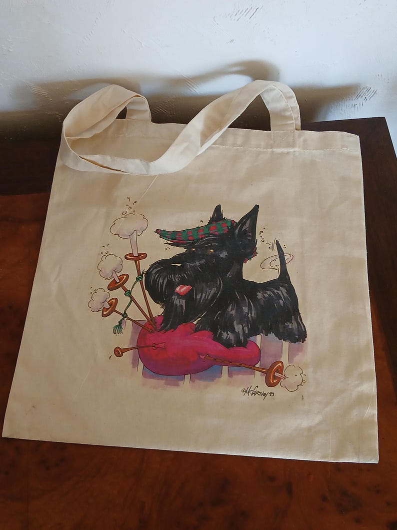 Puede incluir: Una bolsa de mano de lona beige con una ilustraci&oacute;n de dibujos animados de un terrier escoc&eacute;s negro tocando la gaita. El perro lleva un sombrero a cuadros verde y rojo. La bolsa tiene dos asas y forma rectangular. El dise&ntilde;o incluye una gaita roja y humo blanco.