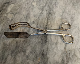 May include: Silver-plated tongs with a spatula-like head. The tongs have ornate handles and are designed for serving food.