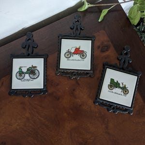 May include: Three vintage car-shaped trivets with black metal frames. The trivets feature illustrations of antique cars with the text "Oldsmobile, 1903", "Duryea Motor Wagon, 1893", and "Cadillac Automobile, 1903".
