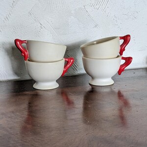 Puede incluir: Cuatro tazas de té pequeñas apiladas con exterior color crema y asas rojas brillantes. Las tazas están colocadas sobre una superficie de madera oscura, con una pared blanca texturizada en el fondo. Las tazas tienen una forma clásica y redondeada.