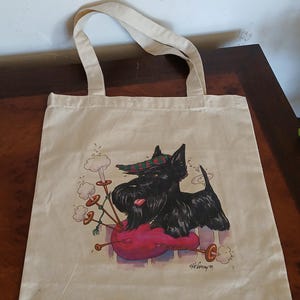 Puede incluir: Un bolso tote beige con una ilustraci&oacute;n de un perro Scottish Terrier negro con un sombrero a cuadros, descansando sobre un coj&iacute;n rosa. El perro est&aacute; al lado de una gaita. El bolso tiene dos asas beige.