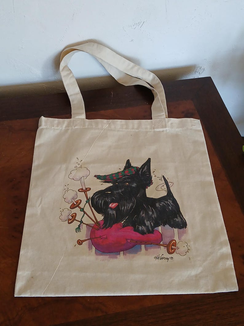 Puede incluir: Un bolso tote beige con una ilustraci&oacute;n de un perro Scottish Terrier negro con un sombrero a cuadros, descansando sobre un coj&iacute;n rosa. El perro est&aacute; al lado de una gaita. El bolso tiene dos asas beige.