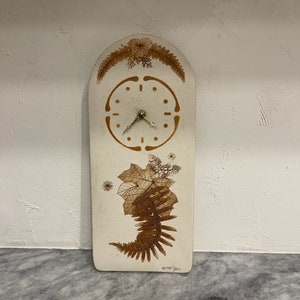 Könnte beinhalten: Eine weiße Keramikuhr mit braunem Farn- und Blattdesign. Das Zifferblatt der Uhr hat einen goldfarbenen Rahmen und Zeiger. Die Uhr ist bogenförmig und trägt unten eine Signatur.