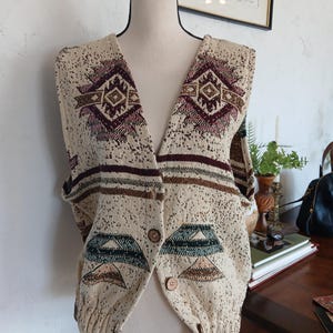 May include: Cream-colored cardigan sweater with a V-neck and button closure. The sweater features geometric patterns in burgundy, brown, and green. The design includes diamond and triangle shapes. The sweater is displayed on a mannequin.
