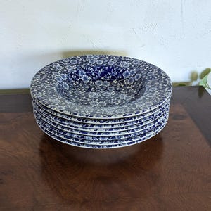 Puede incluir: Una pila de seis platos con estampado floral azul y blanco. Los platos tienen forma de cuenco profundo y un diseño floral detallado. Los platos están apilados sobre una superficie de madera oscura, con una pared blanca al fondo.