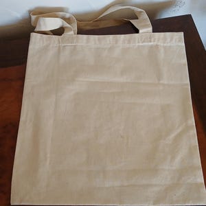 Puede incluir: Una bolsa de lona beige con asas a juego. La bolsa es rectangular y parece estar vac&iacute;a. Las asas est&aacute;n unidas en la parte superior de la bolsa. La bolsa est&aacute; sobre una superficie marr&oacute;n.