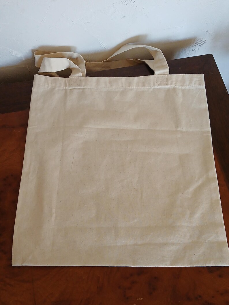 Puede incluir: Una bolsa de lona beige con asas a juego. La bolsa es rectangular y parece estar vac&iacute;a. Las asas est&aacute;n unidas en la parte superior de la bolsa. La bolsa est&aacute; sobre una superficie marr&oacute;n.