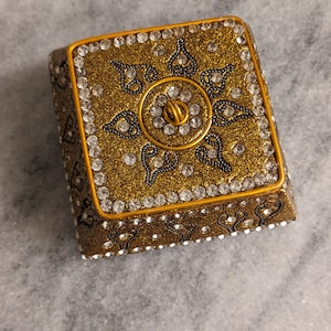 Pode incluir: Uma caixa quadrada dourada com um design decorativo de redemoinhos pretos e strass. A caixa tem um centro elevado com um design circular de strass.