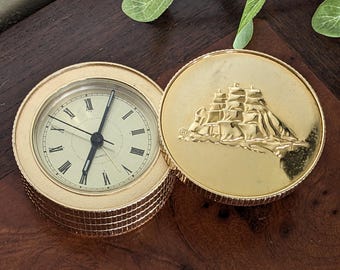 May include: Gold-toned clock with a ship design on the back. The clock face has black hands and Roman numerals. The clock is on a wooden surface.