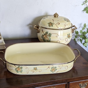 Może przedstawiać: Kremowa emaliowana brytfanna i garnek z dopasowaną pokrywką. Oba elementy mają wzór winorośli i liści w kolorze zielonym i pomarańczowym. Uchwyty i obramowania są w kolorze złotym, a zestaw jest prezentowany na drewnianej powierzchni.