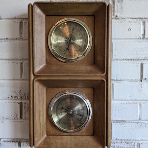 May include: A wooden framed wall mounted barometer and thermometer set. The barometer has a silver face with black markings and the text "RAIN BAROMETER" and "FAIR". The thermometer has a gold face with black markings and the text "THERMOMETER".