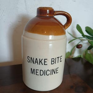 May include: A ceramic jug with a cream-colored body and a brown glazed top section. The jug features a handle, a cork stopper, and the black printed text "SNAKE BITE MEDICINE" on the front. The vessel has a cylindrical shape with a rounded shoulder.