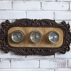 Puede incluir: Un barómetro de pared vintage con marco de madera con tres manómetros analógicos redondos. Los manómetros tienen un acabado dorado y están colocados sobre un fondo beige.