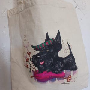 Puede incluir: Bolso tote de lona beige con un Terrier escoc&eacute;s negro con un sombrero a cuadros verde y rojo, tocando una gaita rosa. El perro tiene la lengua fuera. El bolso tiene un asa.
