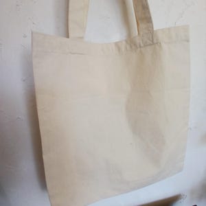 Puede incluir: Un bolso tote de color crema con dos asas a juego. La bolsa est&aacute; hecha de una tela lisa y sin adornos y cuelga contra una pared blanca texturizada. El dise&ntilde;o simple de la bolsa sugiere que es reutilizable y adecuada para transportar varios art&iacute;culos.