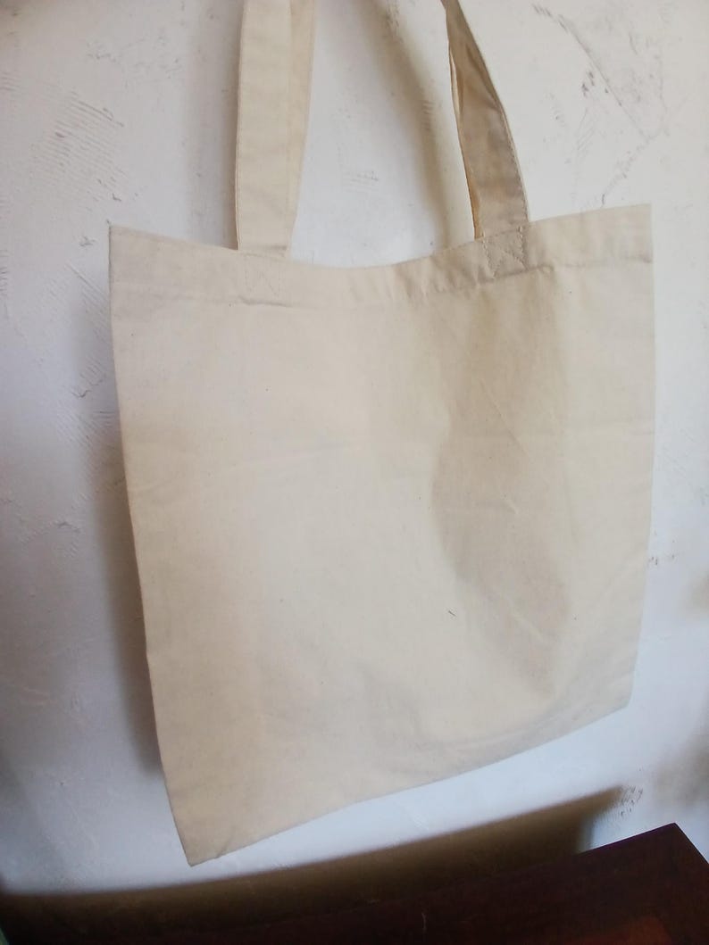 Puede incluir: Un bolso tote de color crema con dos asas a juego. La bolsa est&aacute; hecha de una tela lisa y sin adornos y cuelga contra una pared blanca texturizada. El dise&ntilde;o simple de la bolsa sugiere que es reutilizable y adecuada para transportar varios art&iacute;culos.