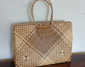 Peut inclure: Un sac à main rectangulaire fabriqué à partir de fibres naturelles tressées dans des tons marron clair et foncé. Le sac présente une fermeture éclair sur le dessus, deux anses circulaires tressées et une patte en V tressée.