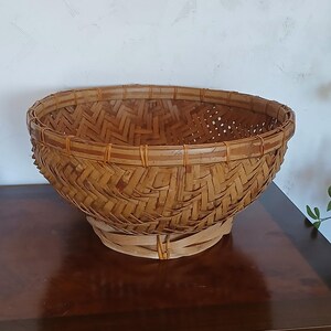 Puede incluir: Una cesta redonda de bambú tejido, con una parte superior ancha y abierta y una base ligeramente ensanchada. La cesta está hecha de tiras de bambú de color marrón claro, intrincadamente tejidas en un patrón de espiga. La cesta está sobre una superficie de madera oscura.