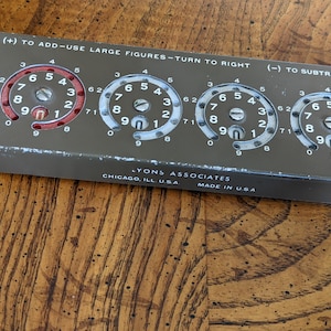 Peut inclure: Une machine à calculer vintage en métal avec quatre cadrans rotatifs. Chaque cadran comporte les chiffres de 0 à 9 et un indicateur rouge ou blanc. La machine comporte des instructions pour l'addition et la soustraction. Le texte indique "(+) TO ADD-USE LARGE FIGURES-TURN TO RIGHT (-) TO SUBTRACT-USE SM". La machine est fabriquée par Lyons Associates à Chicago, Illinois, USA.