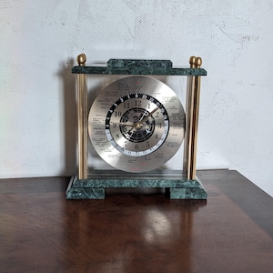 Könnte beinhalten: Eine grüne Marmoruhr mit goldenen Akzenten und einem silbernen Zifferblatt. Das Zifferblatt der Uhr zeigt eine Weltkarte und die Uhrzeit in verschiedenen Städten der Welt an.