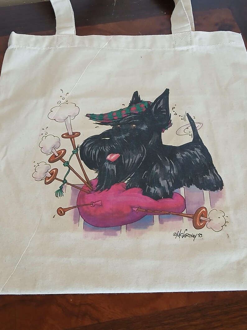 Puede incluir: Bolso tote de color crema con una ilustraci&oacute;n de dibujos animados de un Scottish Terrier negro tocando la gaita. El perro lleva un sombrero a cuadros y descansa sobre una gaita rosa. La gaita tiene detalles en marr&oacute;n y dorado. La firma del artista es visible.