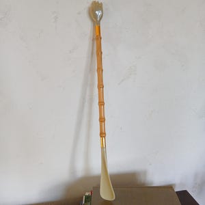 May include: A long-handled shoe horn with a bamboo-style shaft and a hand-shaped top. The shoe horn is beige and gold-toned, with a curved end for easy shoe wear. The handle is made of light brown wood.