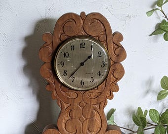 Puede incluir: Un reloj de cuarzo de madera vintage con un diseño tallado y ornamentado. La esfera del reloj es dorada con números y manecillas negras. El reloj es marrón y se encuentra sobre una superficie oscura.
