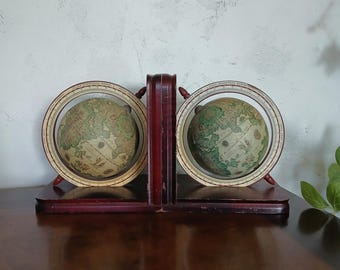 May include: A pair of vintage-style globes mounted on a dark brown wooden base. Each globe is held within a circular frame featuring text labels. The globes display detailed cartographic illustrations in muted earth tones, including greens and tans.