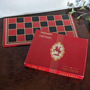 May include: A red box with gold trim and a white knight chess piece on the front. The box says "Tournament Plastic Chessmen" and "E.S. Lowe Co., Inc." The box is on a wooden surface.