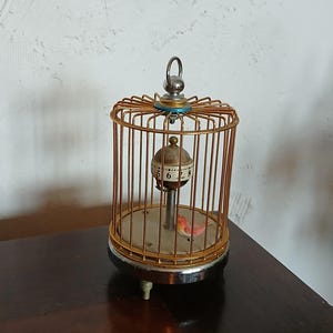 May include: A vintage birdcage music box with a gold-colored wire frame. Inside, a small bird sits on a perch, and a clock face is visible. The base is silver-toned, and the cage has a loop at the top for hanging.