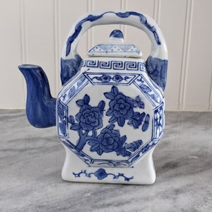 May include: A white and blue ceramic teapot with a floral design. The teapot has an octagonal shape, a curved handle, and a spout. The blue floral pattern decorates the body and lid. The teapot measures approximately 18 cm tall.