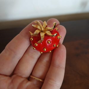 May include: A red strawberry-shaped brooch with gold-colored accents. The brooch features a gold-colored flower on top and small gold-colored dots around the red body. A vintage-style jewelry piece.