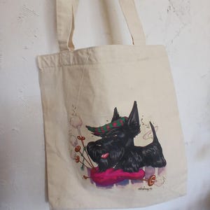 Puede incluir: Bolso tote color crema con un perro Scottish Terrier negro tocando la gaita. El perro lleva un sombrero a cuadros verde y rojo. El bolso tiene dos correas color crema y cuelga de una pared blanca texturizada.