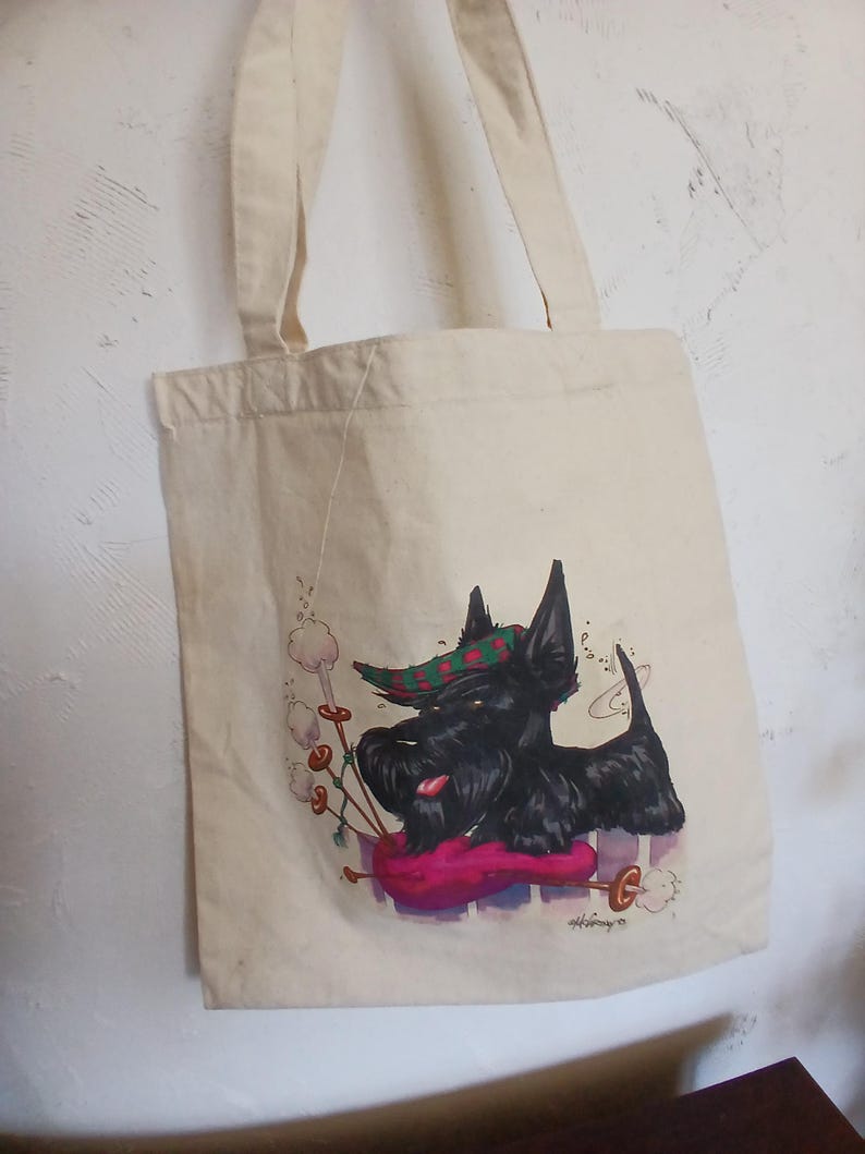 Puede incluir: Bolso tote color crema con un perro Scottish Terrier negro tocando la gaita. El perro lleva un sombrero a cuadros verde y rojo. El bolso tiene dos correas color crema y cuelga de una pared blanca texturizada.