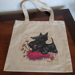 Puede incluir: Un bolso tote beige con una ilustraci&oacute;n de dibujos animados de un perro Scottish Terrier negro tocando la gaita. El perro lleva un sombrero a cuadros y descansa sobre una gaita rosa. La firma del artista est&aacute; en la esquina inferior derecha.
