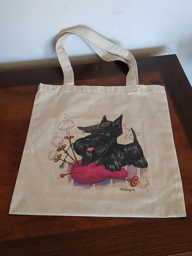 Puede incluir: Un bolso tote beige con una ilustraci&oacute;n de dibujos animados de un perro Scottish Terrier negro tocando la gaita. El perro lleva un sombrero a cuadros y descansa sobre una gaita rosa. La firma del artista est&aacute; en la esquina inferior derecha.